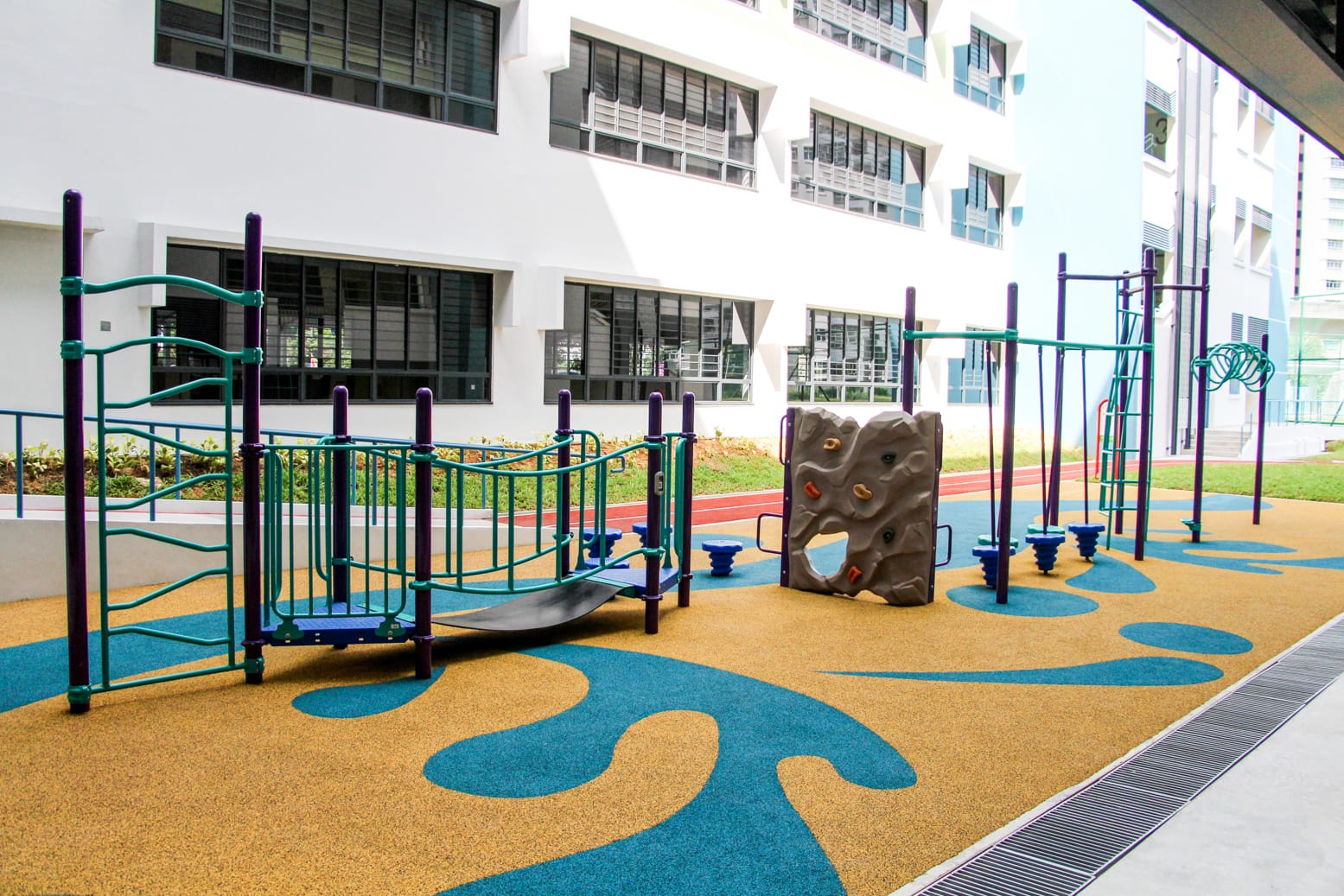 Outdoor playground with climbing frames and a rock wall on teal and tan rubber flooring beside a white building.